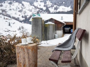 A bench with a wooden backrest leaning against a wall, a wooden stump with two white cups and saucers, and a view of a snowy mountain landscape with buildings in the background.