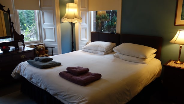 Una cama bien hecha con almohadas y toallas dobladas, una lámpara de noche, un oso de peluche en una mesita de noche y una ventana con vistas a la vegetación.
