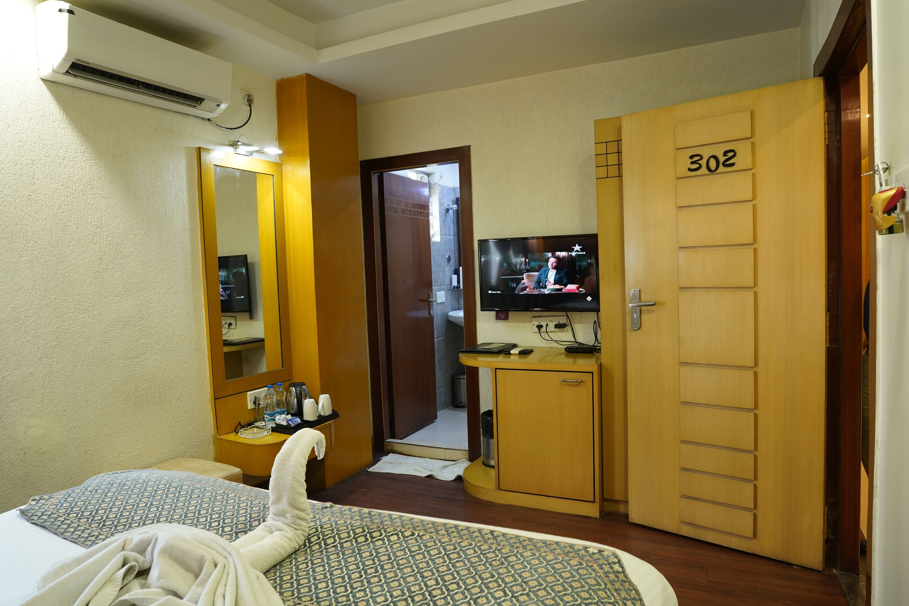 A hotel room with a bed, a TV, a bathroom, and a wooden door with the number 302.