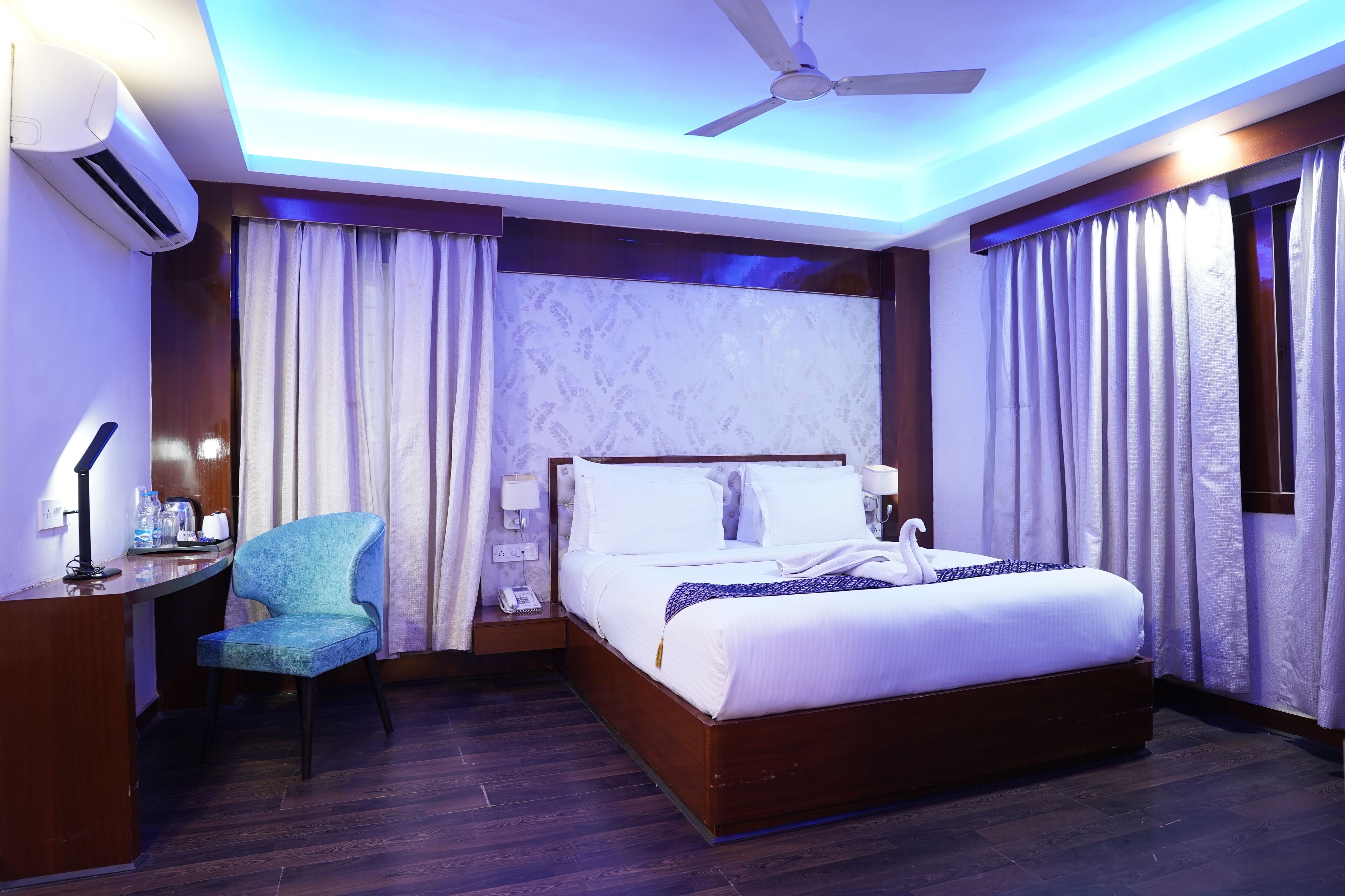 A modern hotel room with a wooden bed, a desk with a chair, a ceiling fan, and an air conditioning unit.