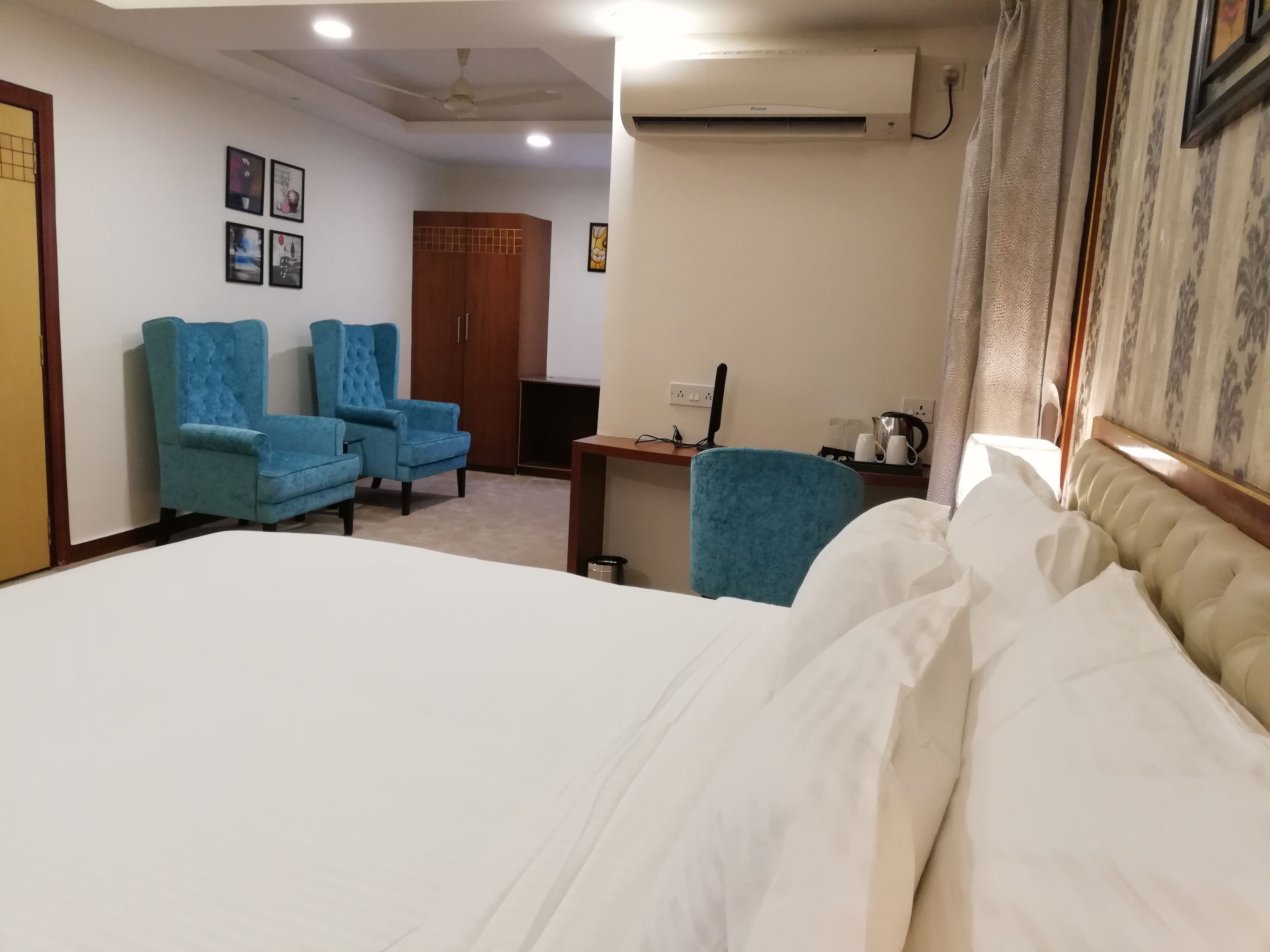 Een hotelkamer met een bed, twee blauwe fauteuils, een houten garderobe, een bureau met een computer en een airconditioner.