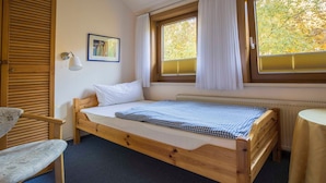 Ein Einzelbett mit karierter Decke, ein Nachttisch, ein Stuhl, ein Fenster mit Vorhängen und eine Heizung.