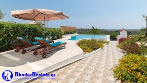 Zona junto a la piscina con sillas de descanso y una sombrilla, rodeada de un jardín ajardinado y vistas al paisaje.