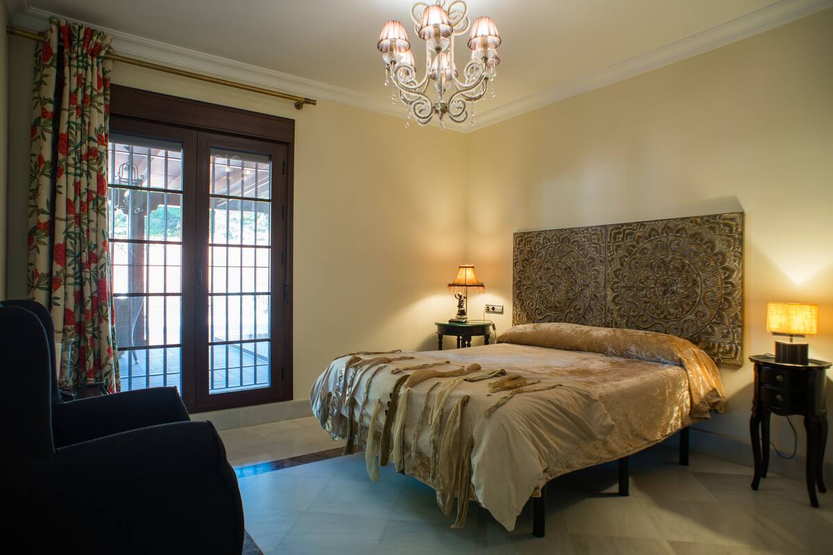 A bedroom with a large bed, a chandelier, a patterned headboard, and a window with curtains.