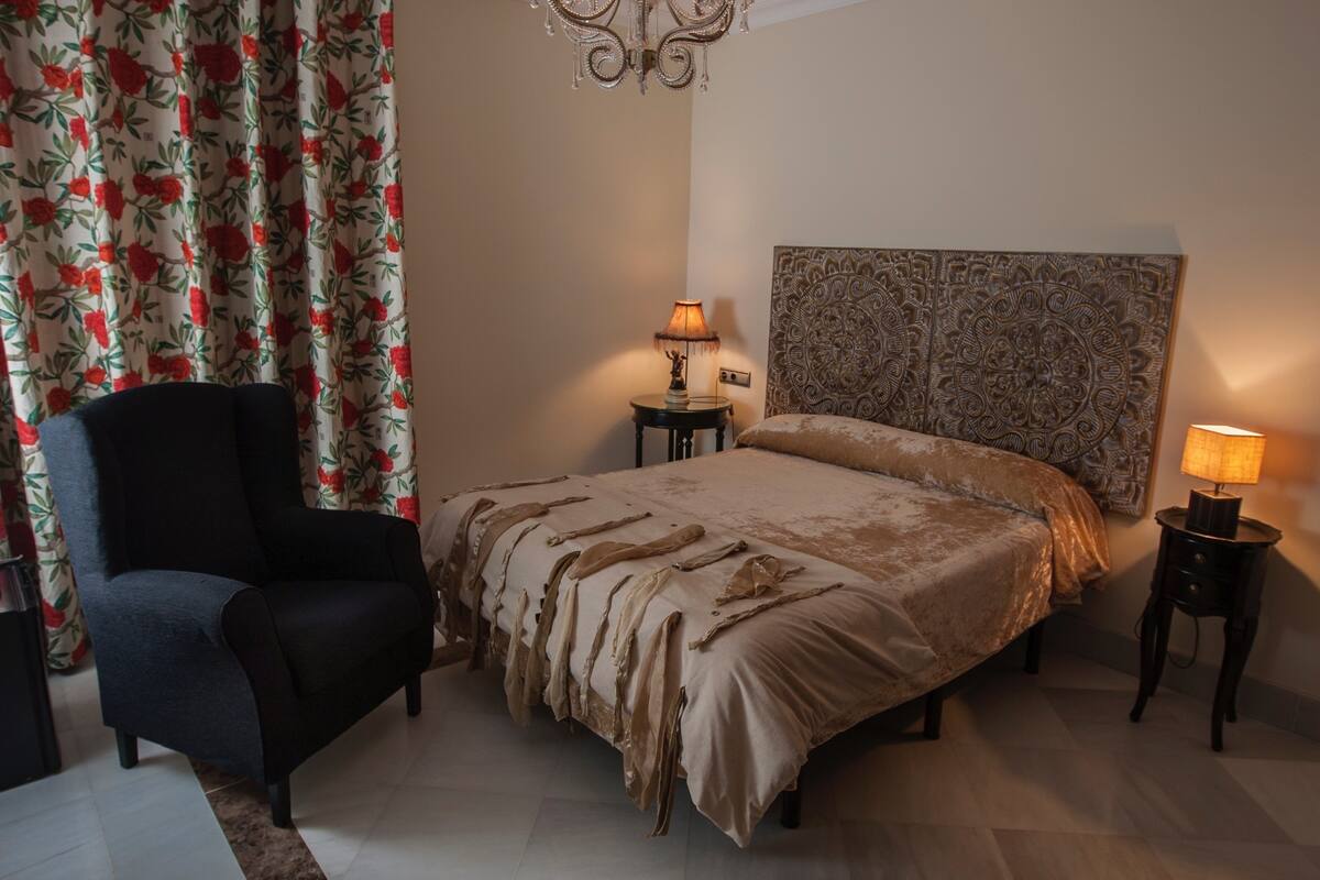 A bedroom with a bed, a chair, two bedside tables with lamps, floral curtains, and a decorative headboard.