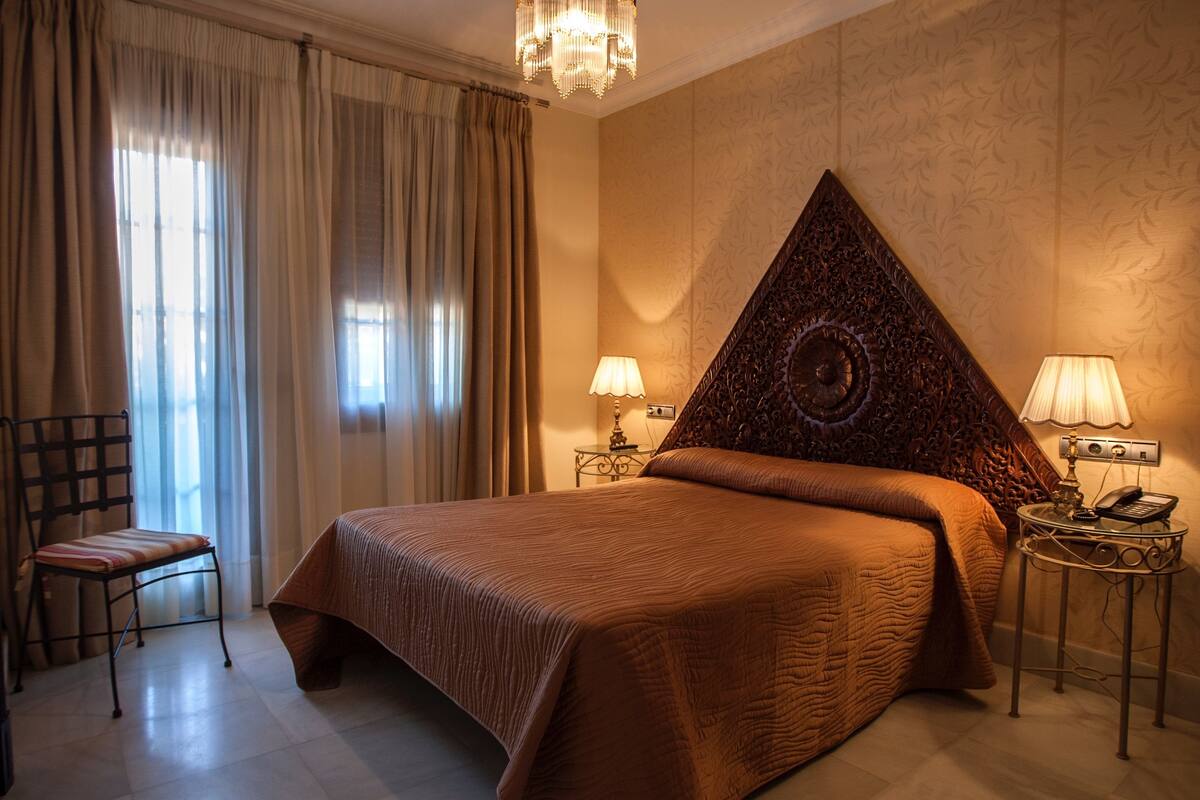 A hotel room with a large bed, two bedside tables with lamps, a chair, and a large window with curtains.