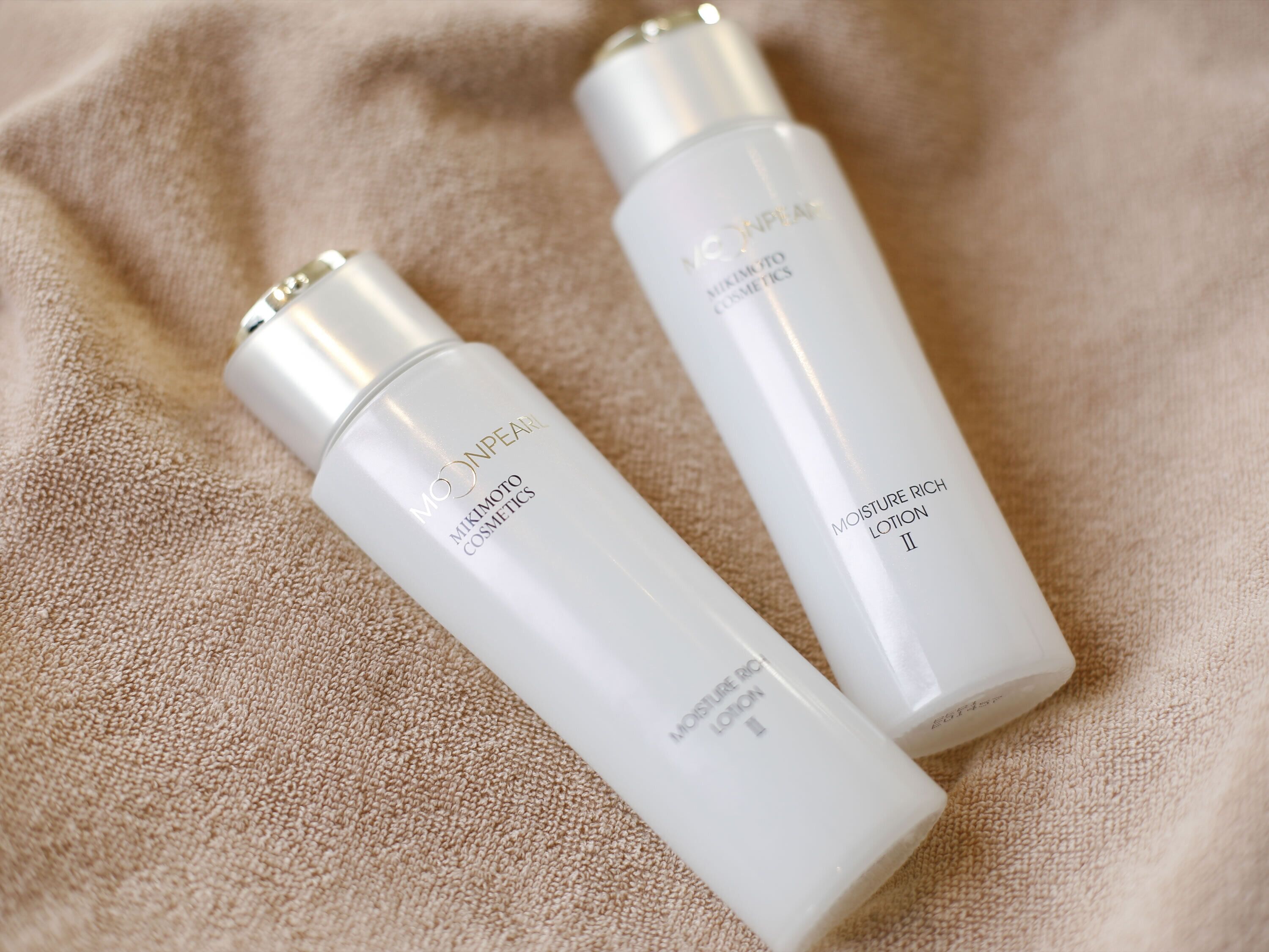 Two bottles of MIKIMOTO COSMETICS on a beige textured surface.