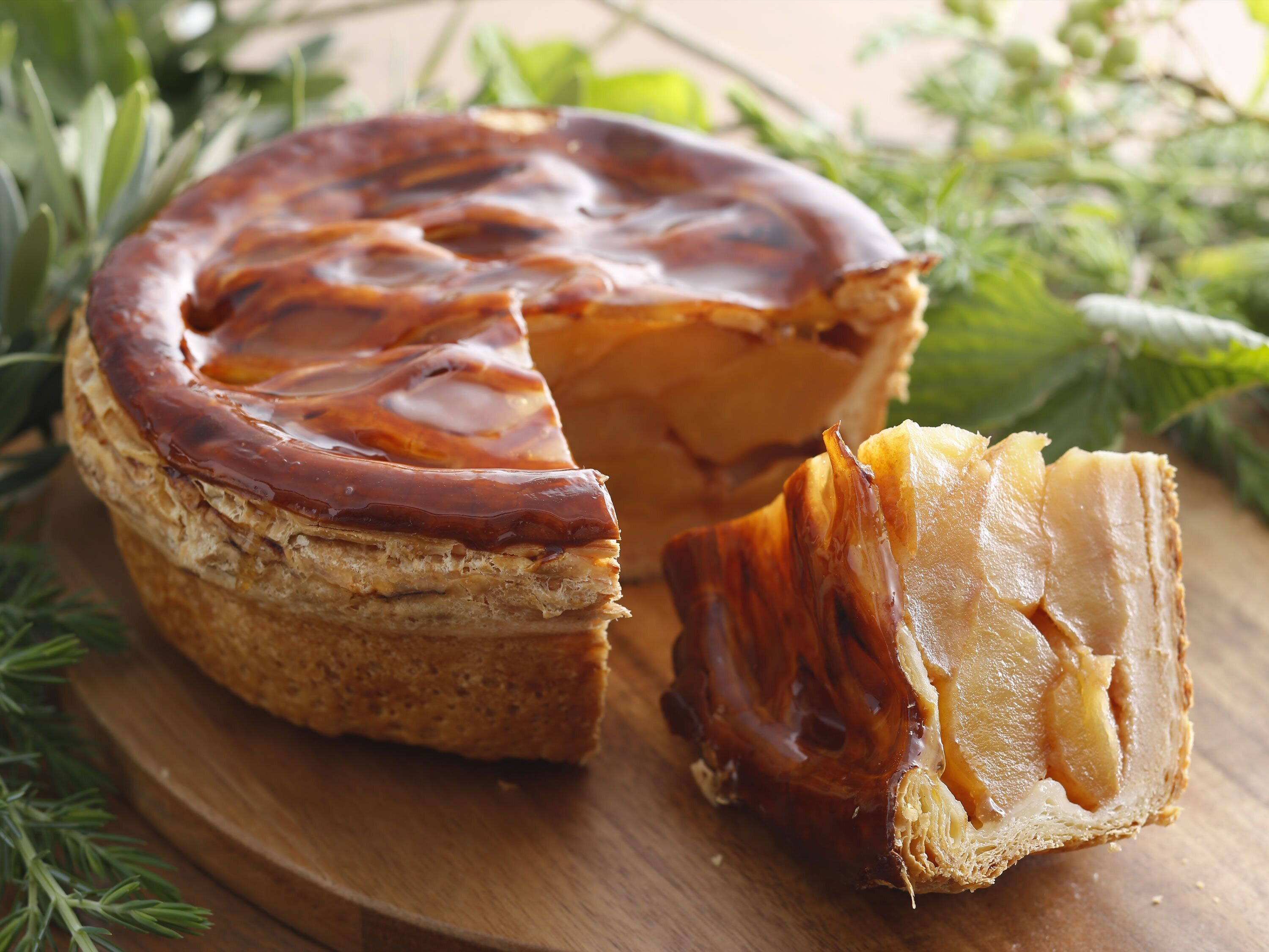 A round pastry with a caramelized top, partially cut, on a wooden board with greenery in the background.