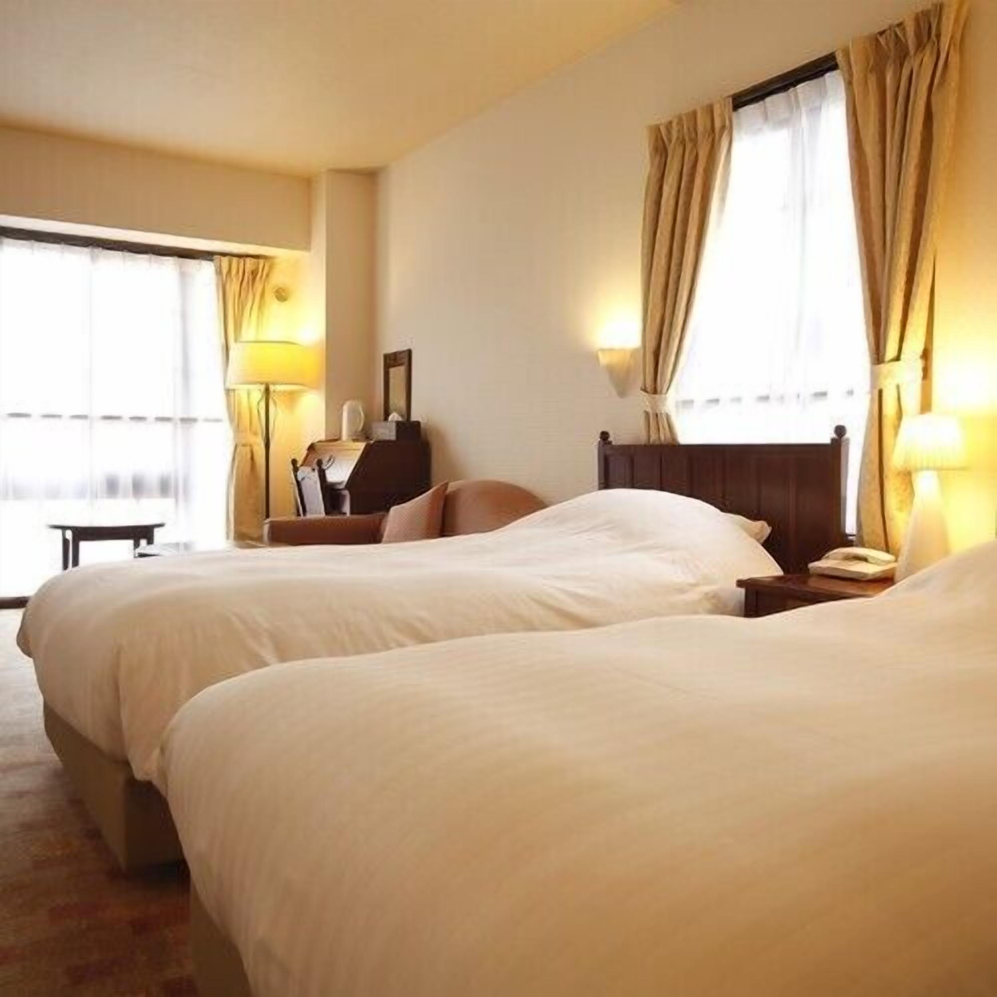 Una habitación de hotel con dos camas, un escritorio, una silla, una lámpara y una ventana con cortinas.