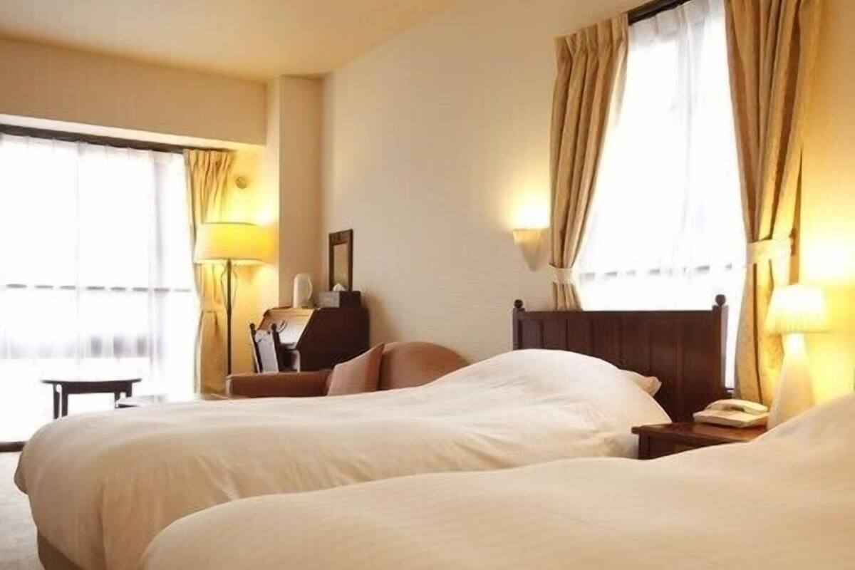 A hotel room with two beds, a desk, a chair, a lamp, and a window with curtains.