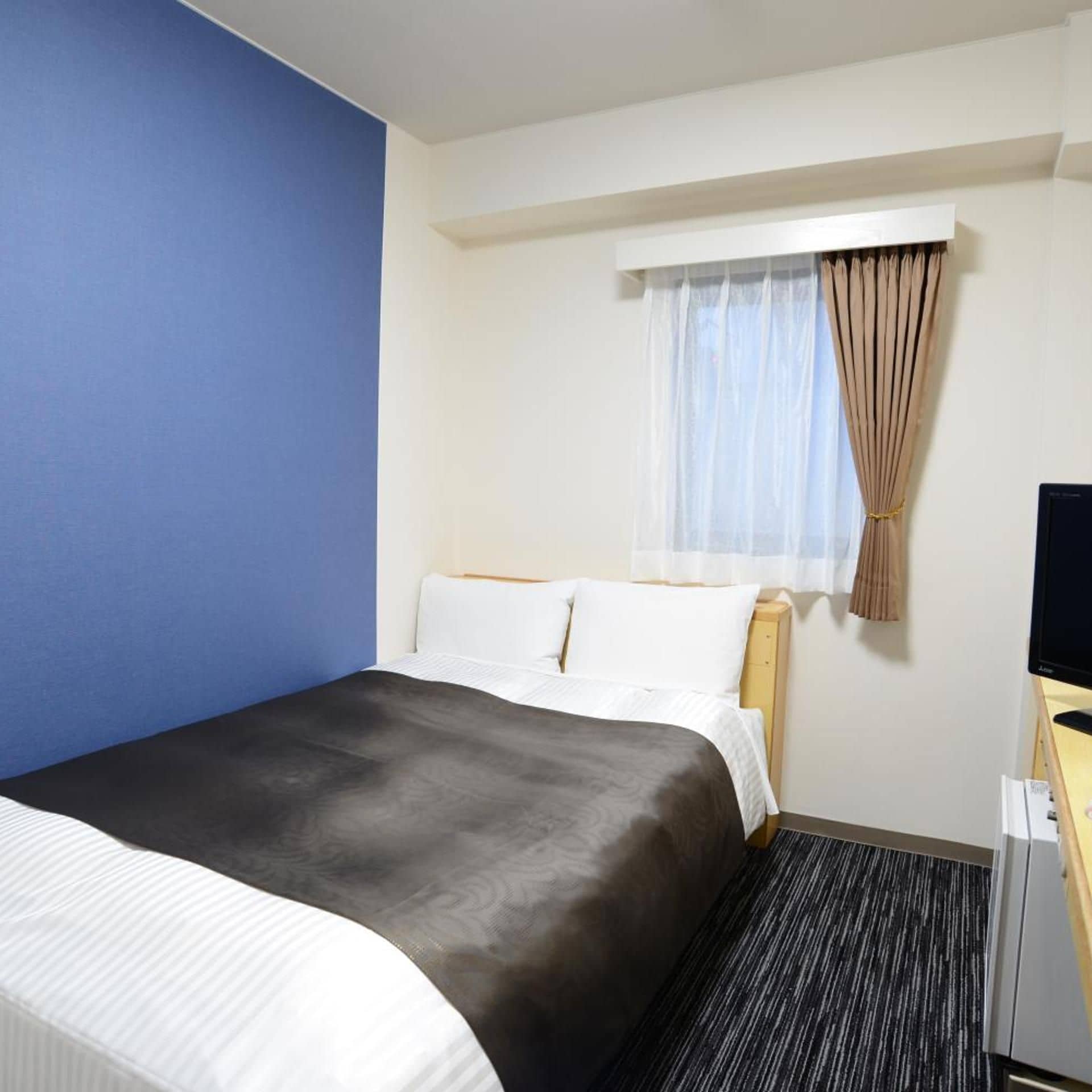 A hotel room with a bed, a television, a window with curtains, and a desk.