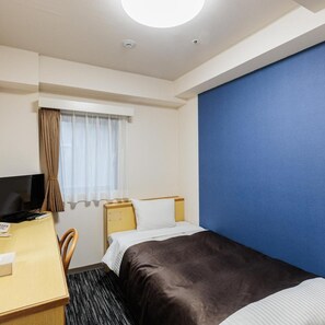A hotel room with a bed, a desk with a computer, a window with curtains, and a blue wall.
