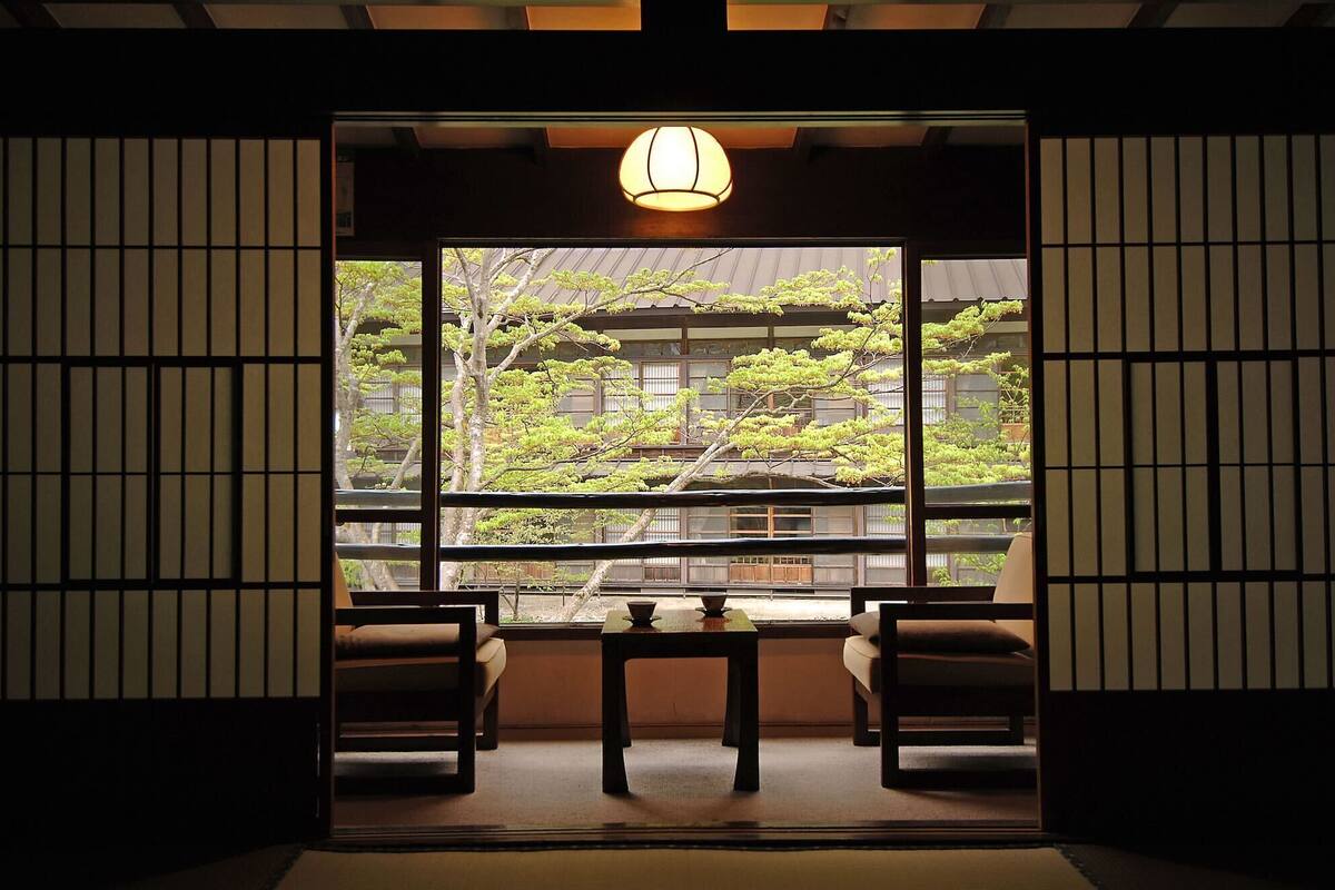 A traditional Japanese room with tatami flooring, a low table, two chairs, and a large window with a view of a building and trees.