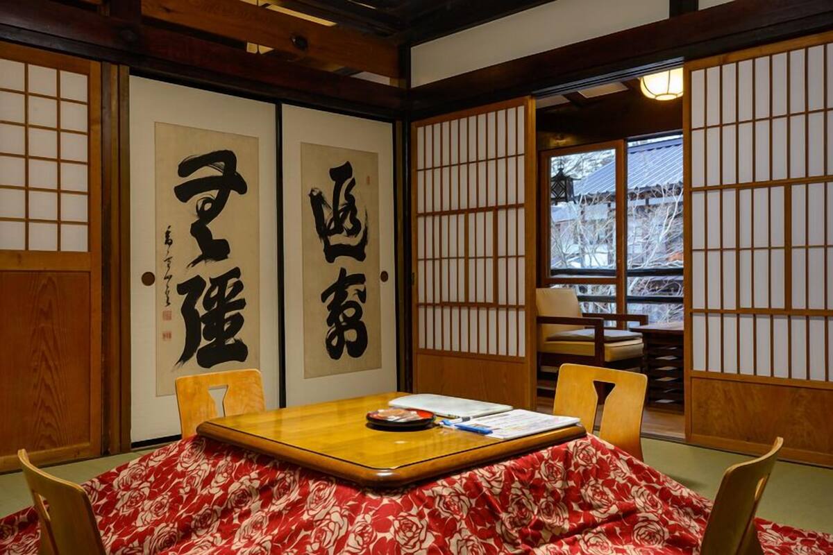 A traditional Japanese room with tatami flooring, a low table with a red floral tablecloth, and sliding doors with calligraphy.