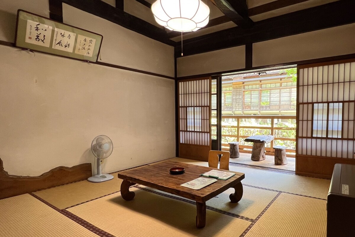 Ein traditioneller japanischer Raum mit Tatami-Boden, einem niedrigen Holztisch, einem Fächer und Schiebetüren zu einem Außenbereich.