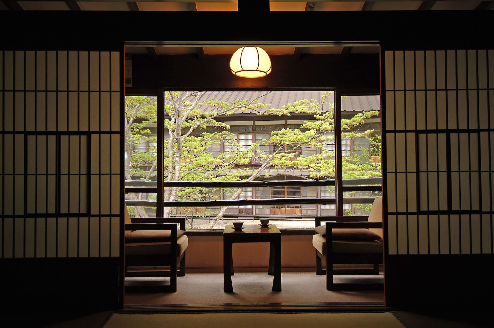 Une pièce de style japonais traditionnel, avec un sol recouvert de tatamis, une table basse, deux chaises et une grande fenêtre donnant sur un bâtiment et des arbres.
