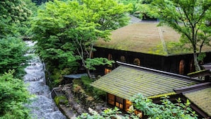 Un edificio japonés tradicional con tejado de tejas, rodeado de abundante vegetación y un pequeño arroyo.