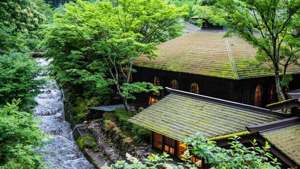 Ein traditionelles japanisches Gebäude mit einem Ziegeldach, umgeben von üppiger Vegetation und einem kleinen Bach.