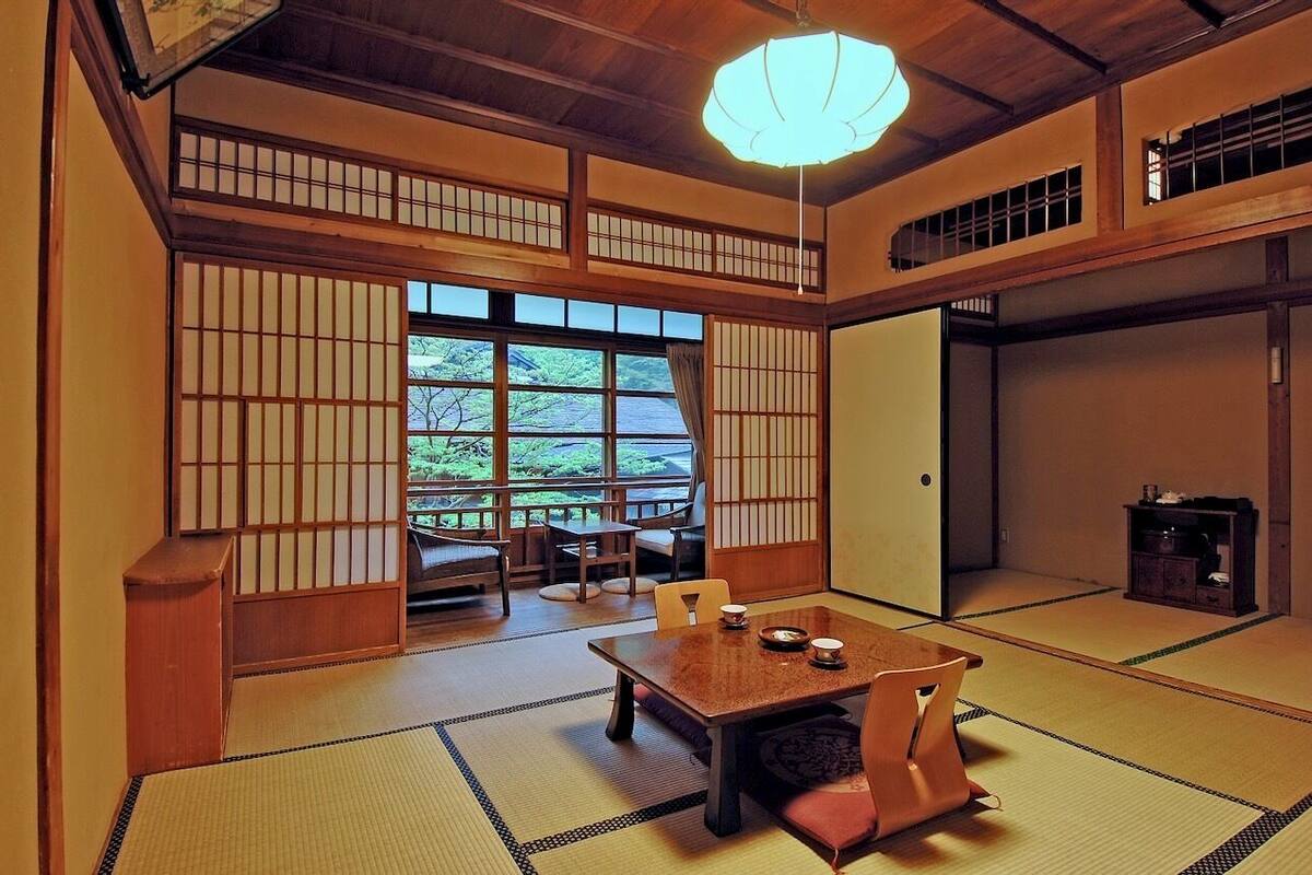 A traditional Japanese room with tatami flooring, a low table with cushions, sliding doors, and a view of greenery outside.