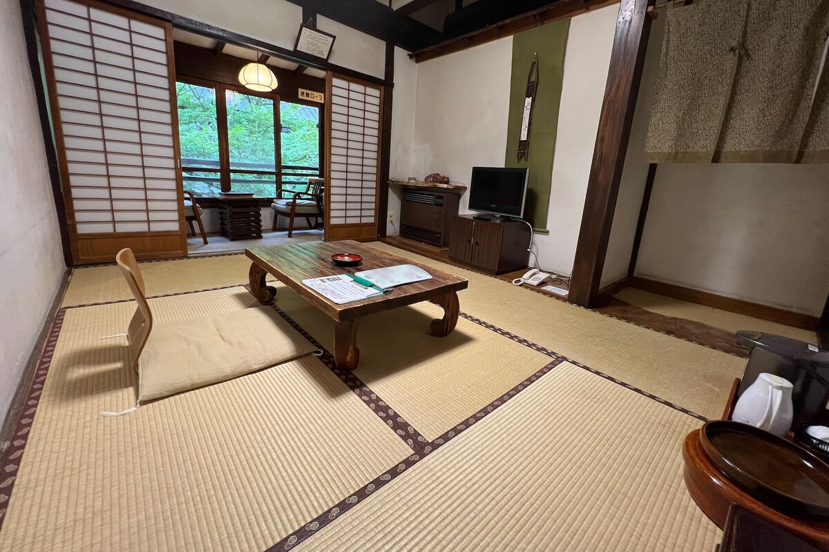 Una habitación japonesa tradicional con suelo de tatami, una mesa baja con cojín, un televisor y una puerta corredera que da a vistas de follaje.