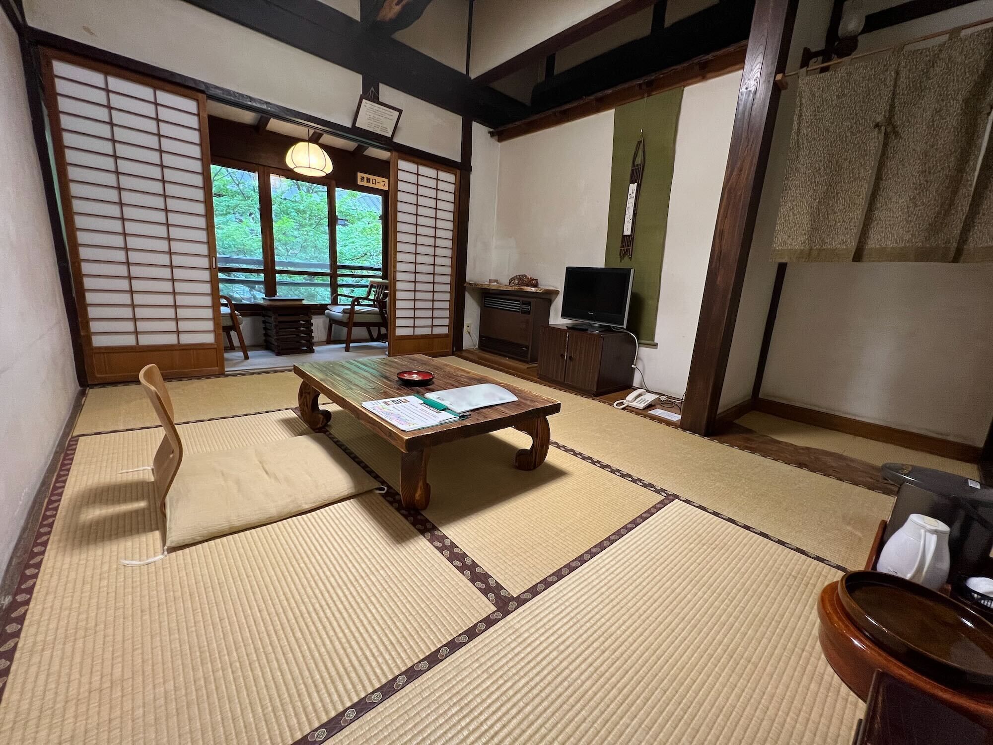 Una habitación japonesa tradicional con suelo de tatami, una mesa baja con cojín, un televisor y una puerta corredera que da a vistas de follaje.