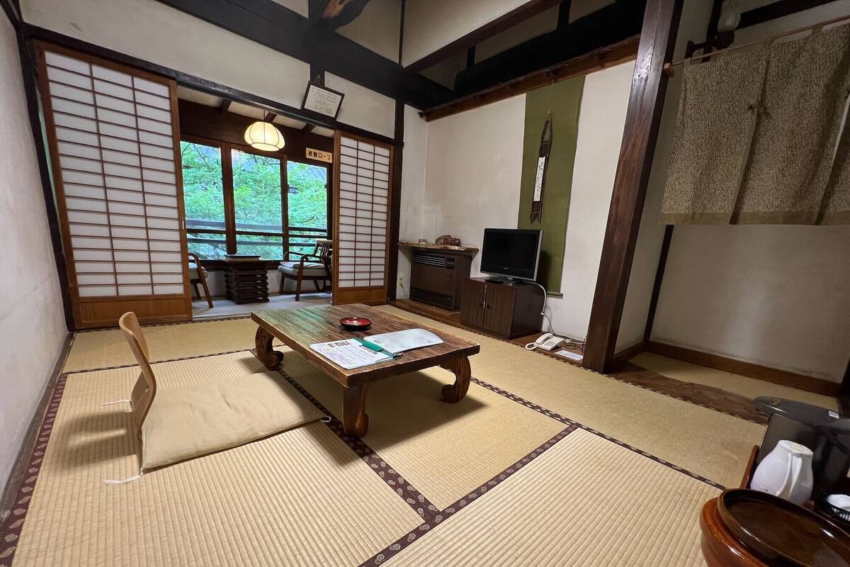 A traditional Japanese room with tatami flooring, a low table with a cushion, a television, and a sliding door leading to a view of greenery.