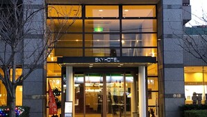 Der Eingang eines Hotels mit Glasfassade, einem Schild mit der Aufschrift "SKYHOTEL" und einem einsehbaren Eingangsbereich mit Stufen, die zur Tür führen.