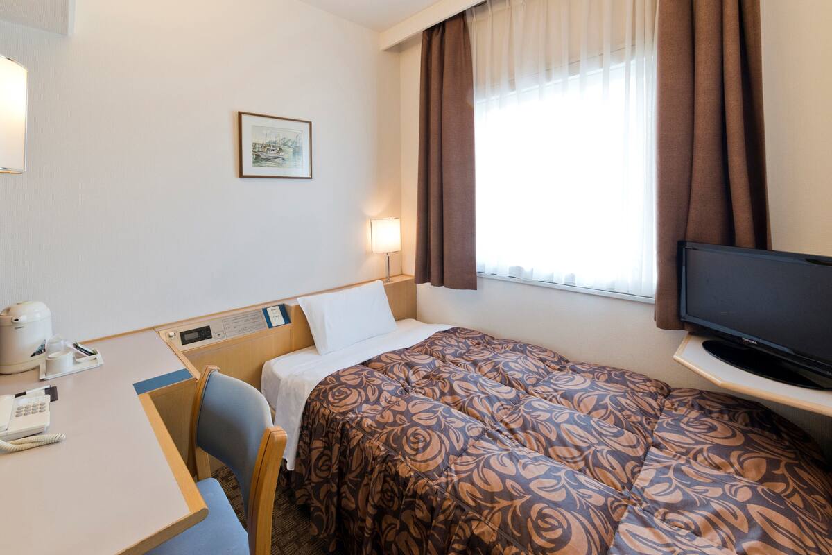 A hotel room with a bed, a desk with a telephone, a television, and a window with curtains.