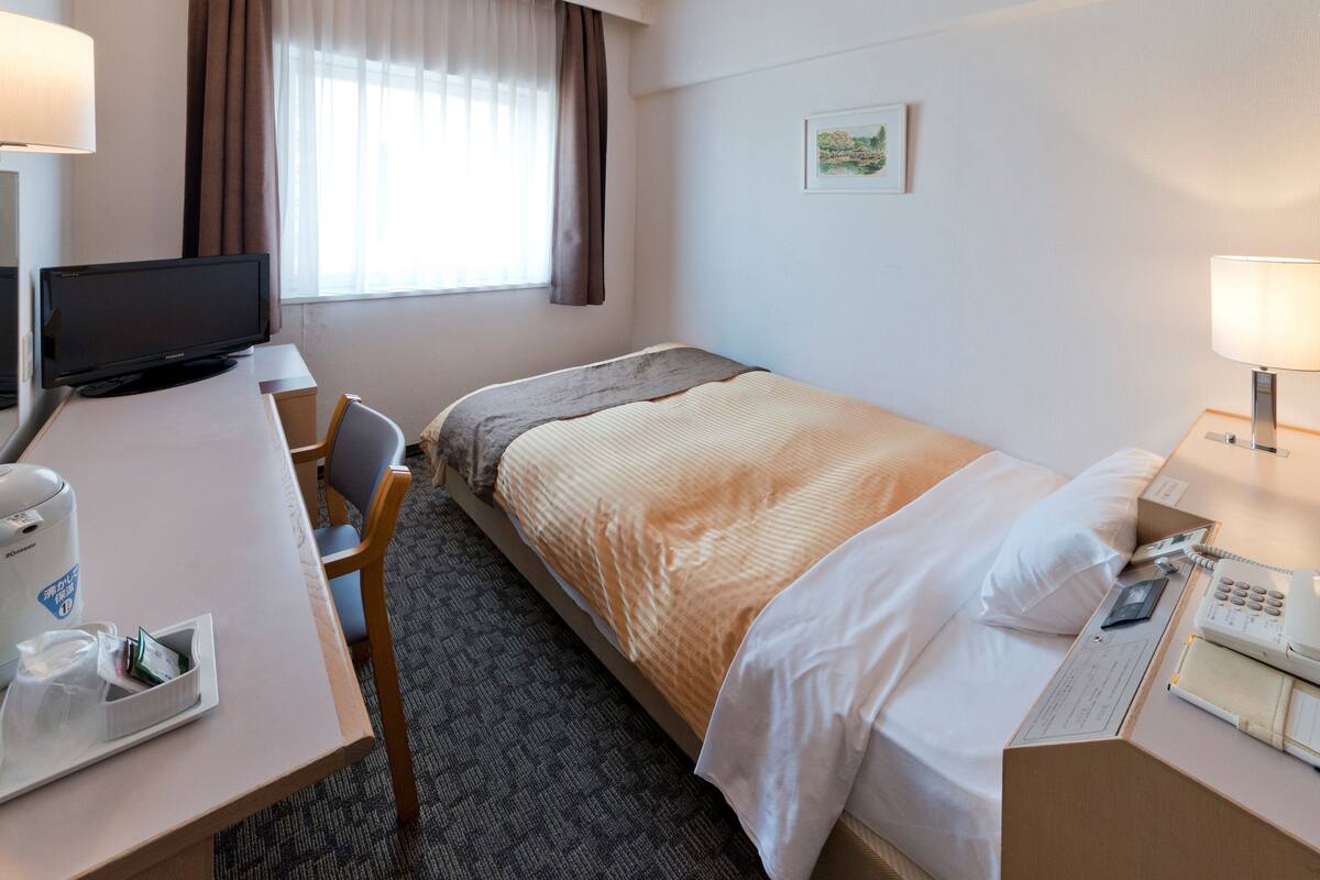A hotel room with a bed, a desk with a computer, a chair, a telephone, and a window with curtains.