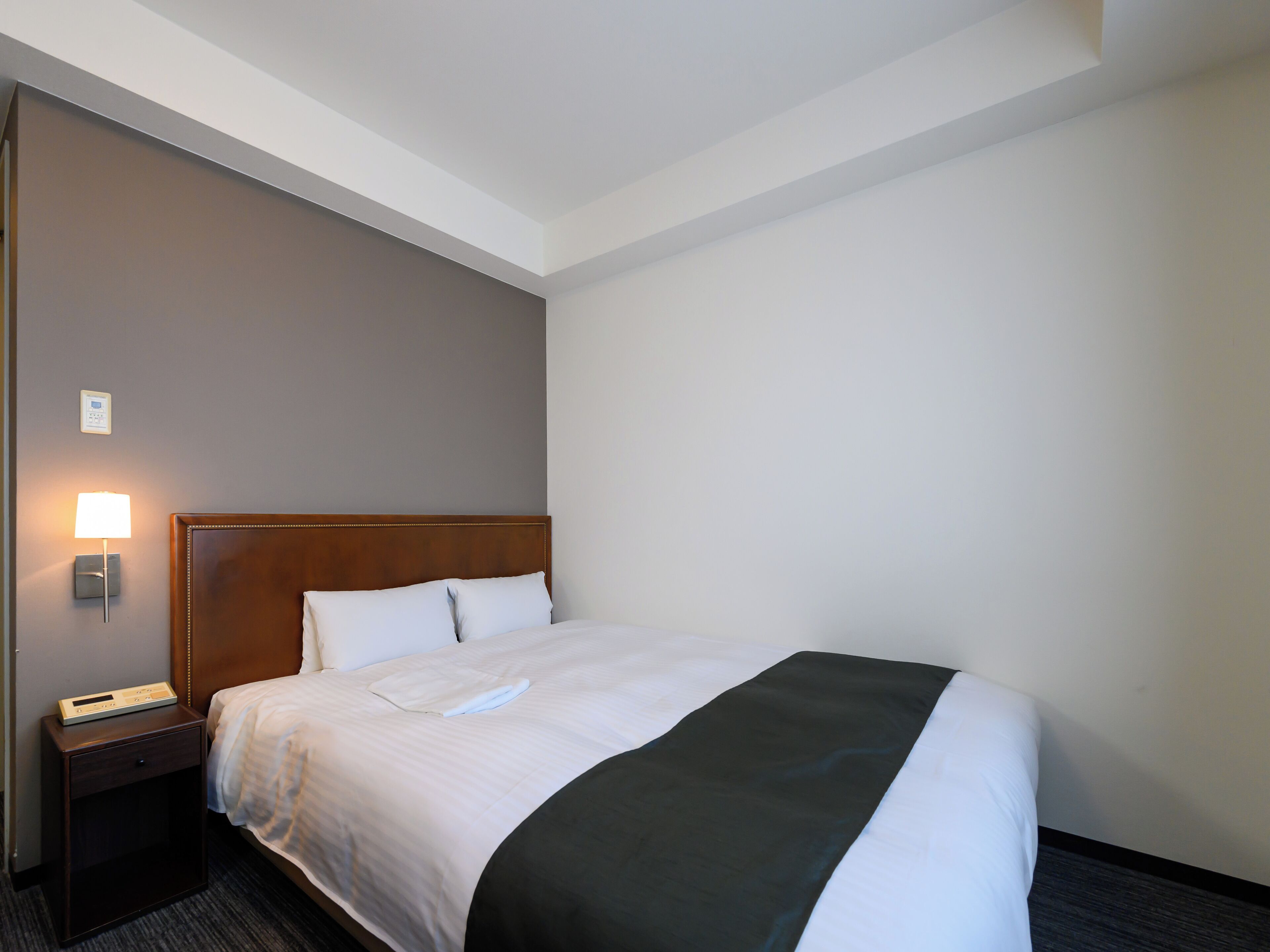 Una habitación de hotel con una cama bien hecha, una mesita de noche con lámpara y un panel de control en la pared.
