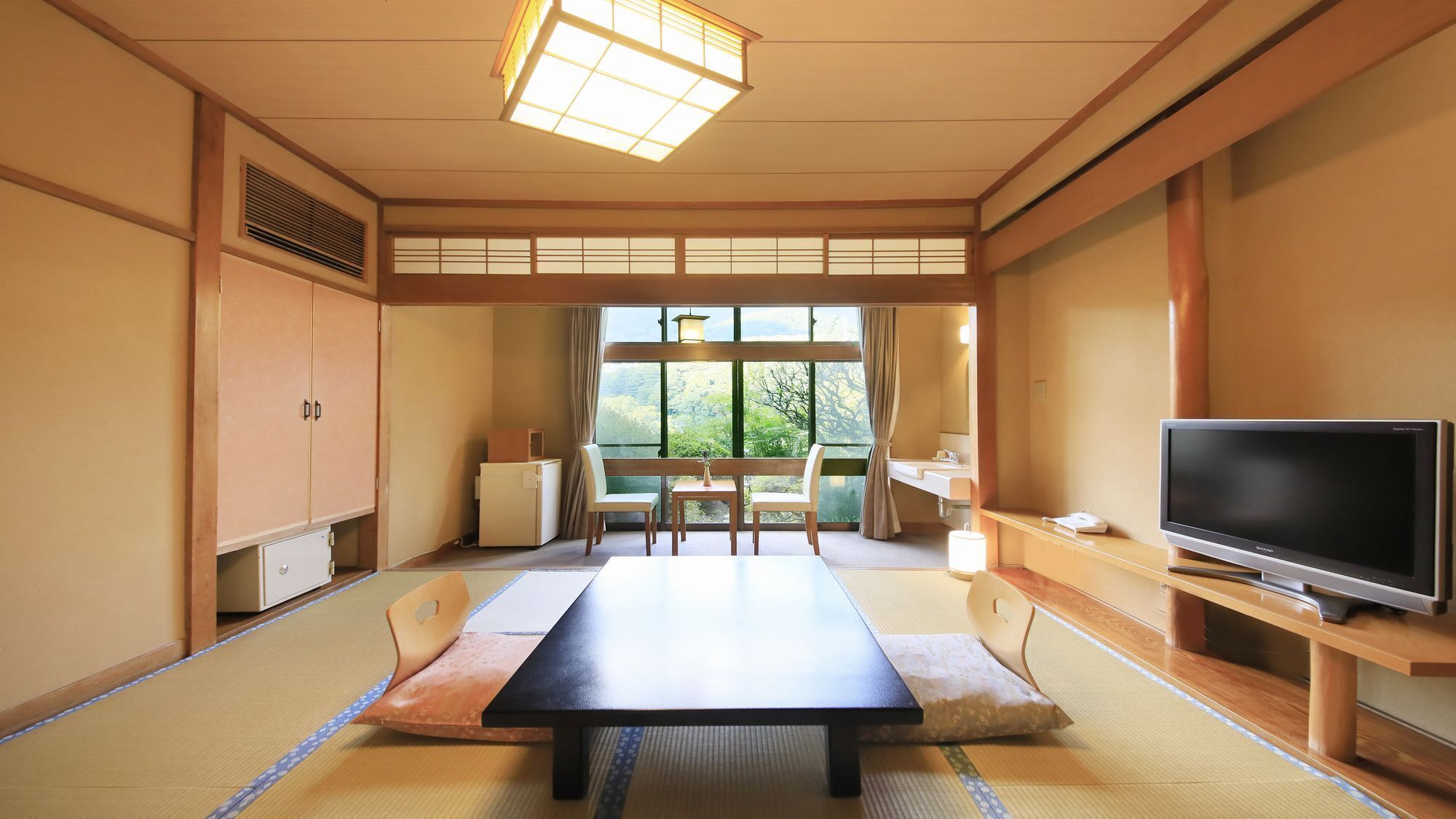 A traditional Japanese room with tatami flooring, a low table, a television, and a view of greenery outside.