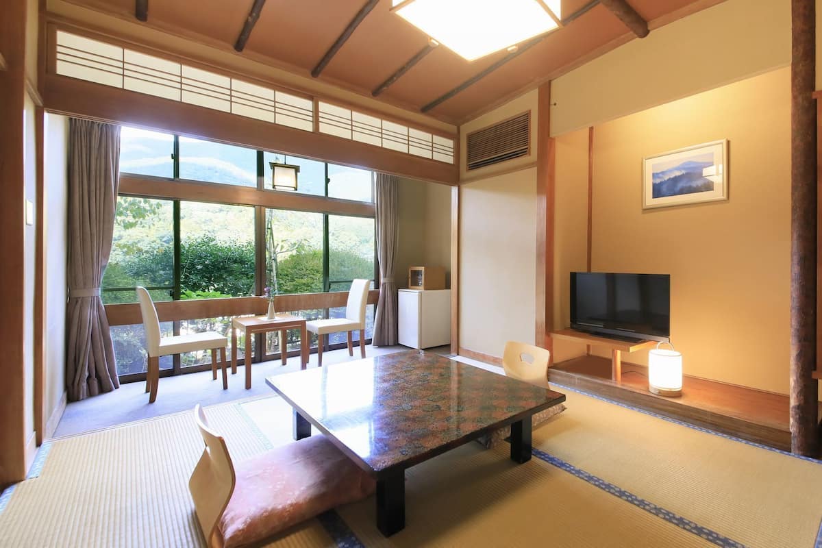 A traditional Japanese-style room with tatami flooring, a low table, a TV, and a view of greenery outside.