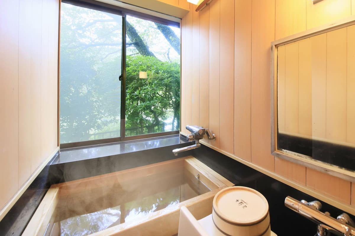 A traditional Japanese-style bathroom with a large soaking tub, a wooden sliding door, and a view of greenery outside.