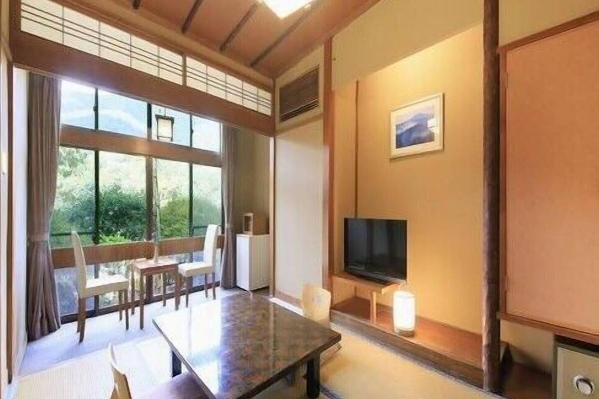 A traditional Japanese room with tatami flooring, a low table, a sofa, a TV, and a window with a view of greenery.