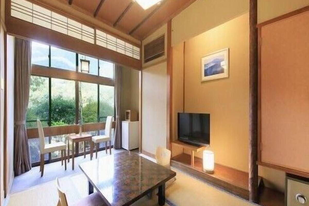 A traditional Japanese room with tatami flooring, a low table, a bench, a television, and a window with a view of greenery.