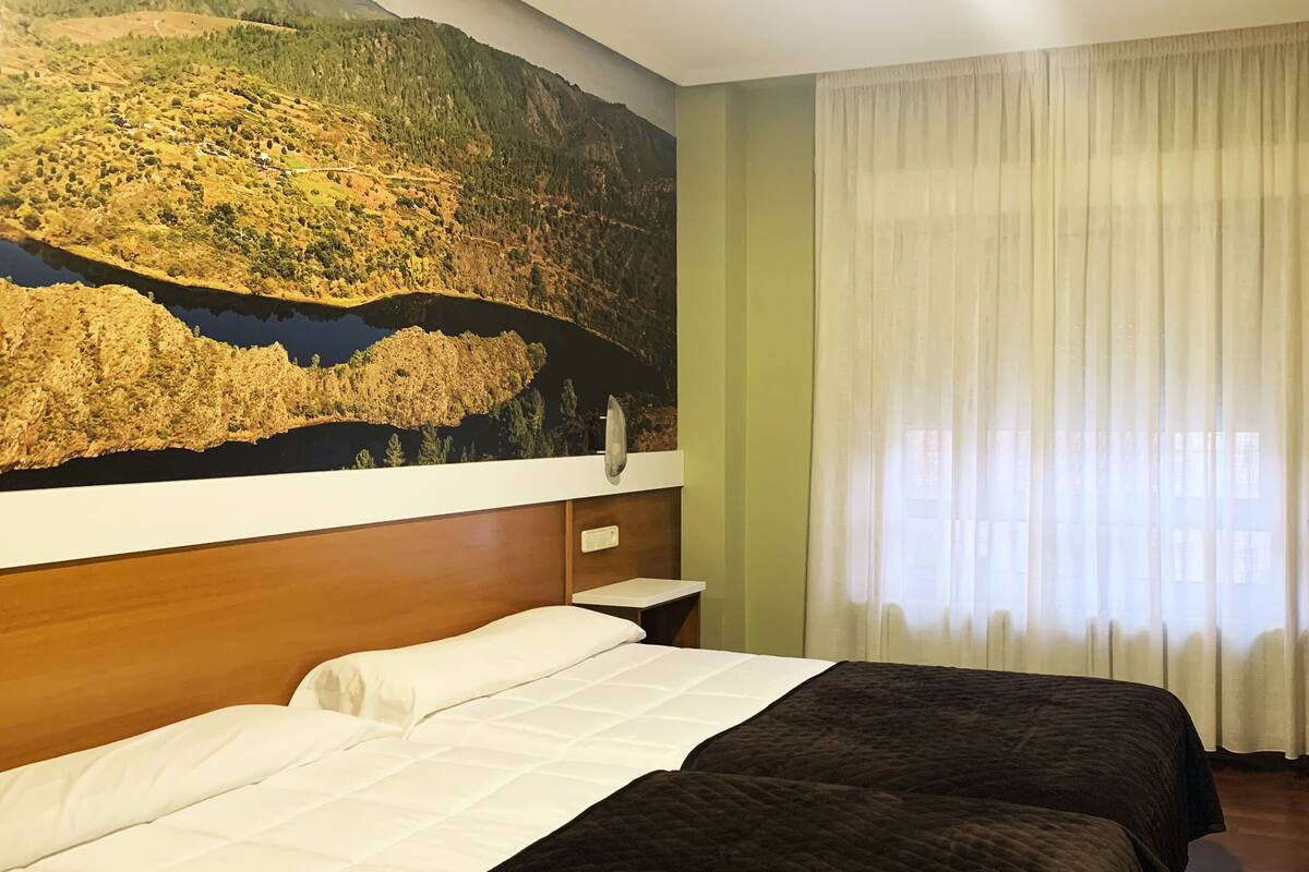 Une chambre d’hôtel avec un grand lit, une table de chevet, un téléviseur fixé au mur et une fenêtre avec des rideaux.