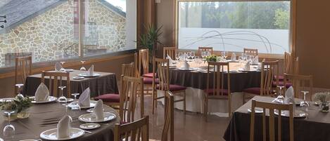 A restaurant with round tables set for dining, large windows with a view of the outside, and a stone wall in the background.