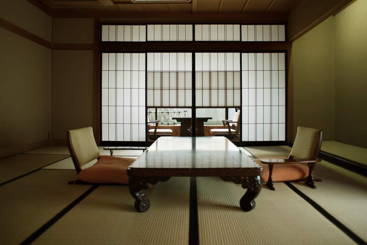 A tatami mat room with a low table and two chairs, large shoji screens, and a view through the screens.