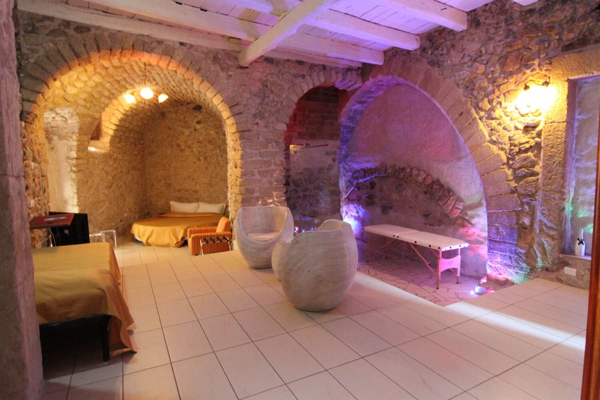 A stone-walled room with a bed, two chairs, a bench, and a table.