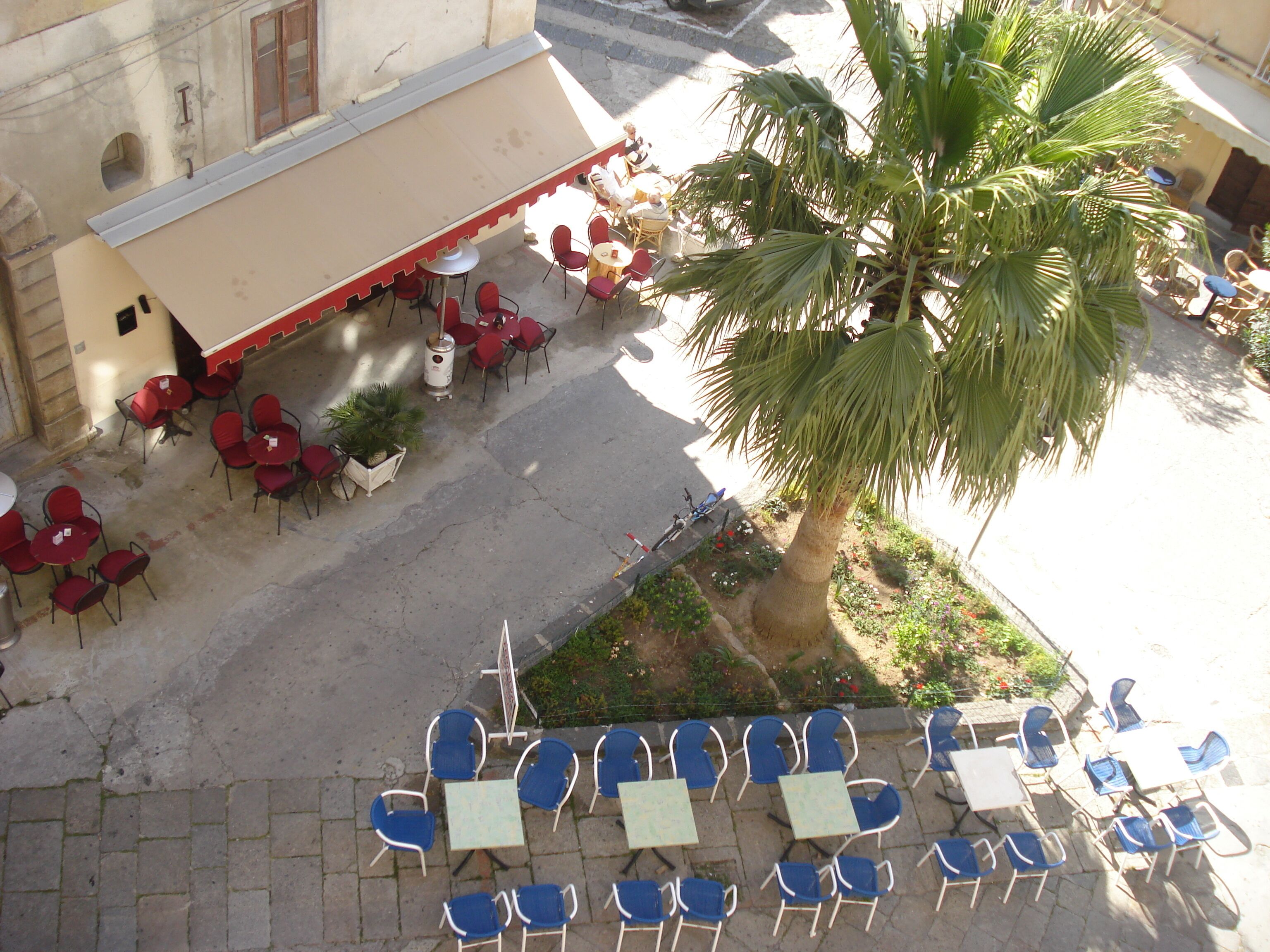 An outdoor seating area with red and blue chairs and tables, a palm tree, and a building with a beige awning.