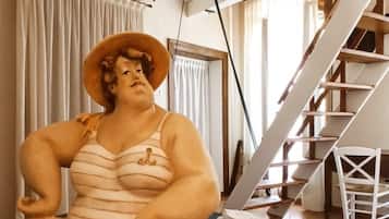 Eine Statue einer Person, die auf einem Tisch in einem Raum sitzt. Im Hintergrund sind eine Treppe und ein Stuhl zu sehen.