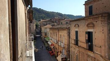 Une rue étroite bordée de bâtiments des deux côtés, un ciel dégagé et un paysage vallonné à l’horizon.