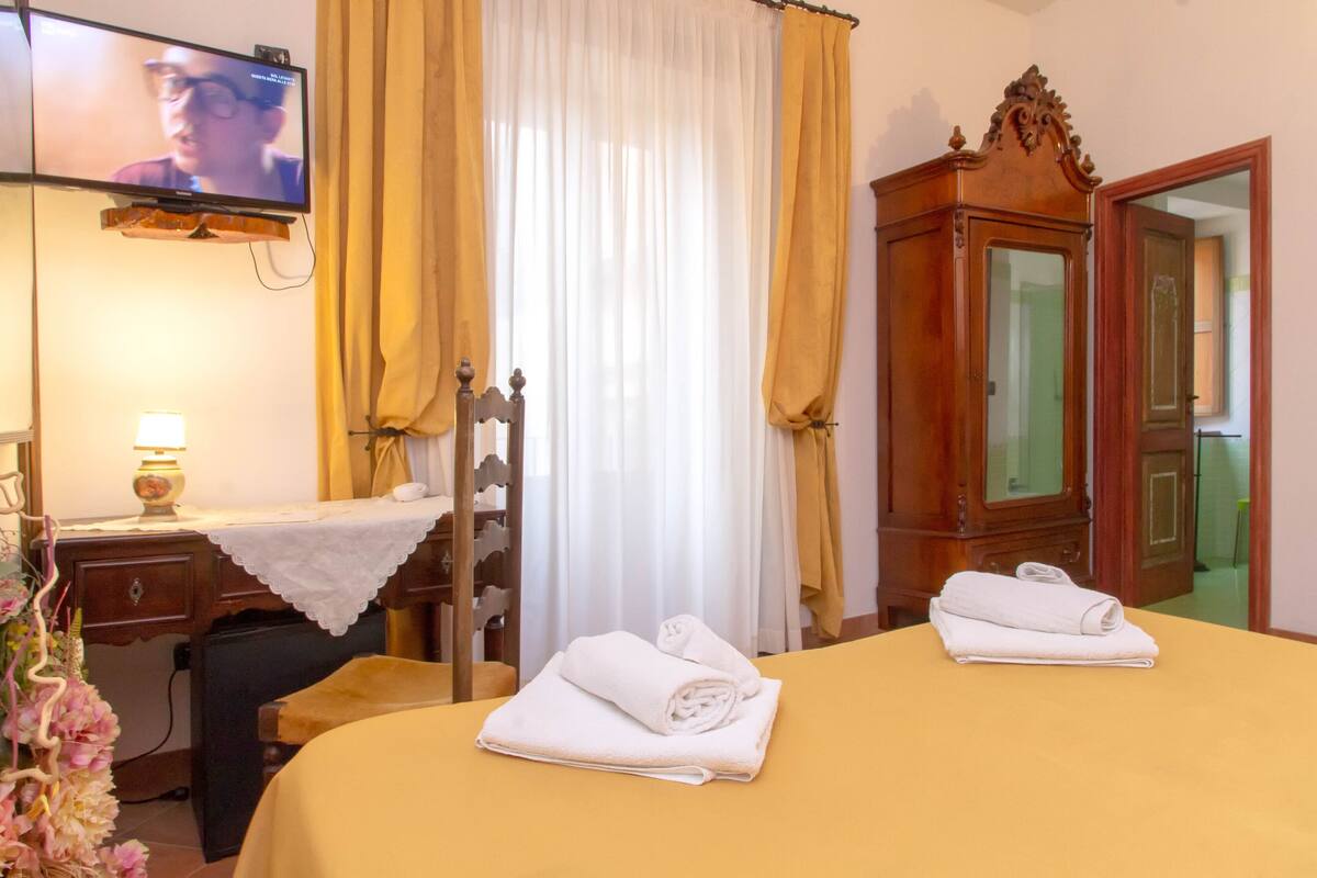 A hotel room with a bed, a desk with a lamp, a television, and a wooden wardrobe.