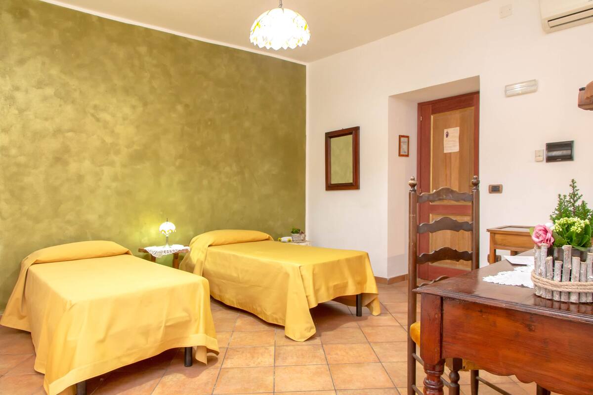 A room with two beds covered in yellow sheets, a wooden nightstand with a lamp, a wooden dresser, and a door with a mirror.