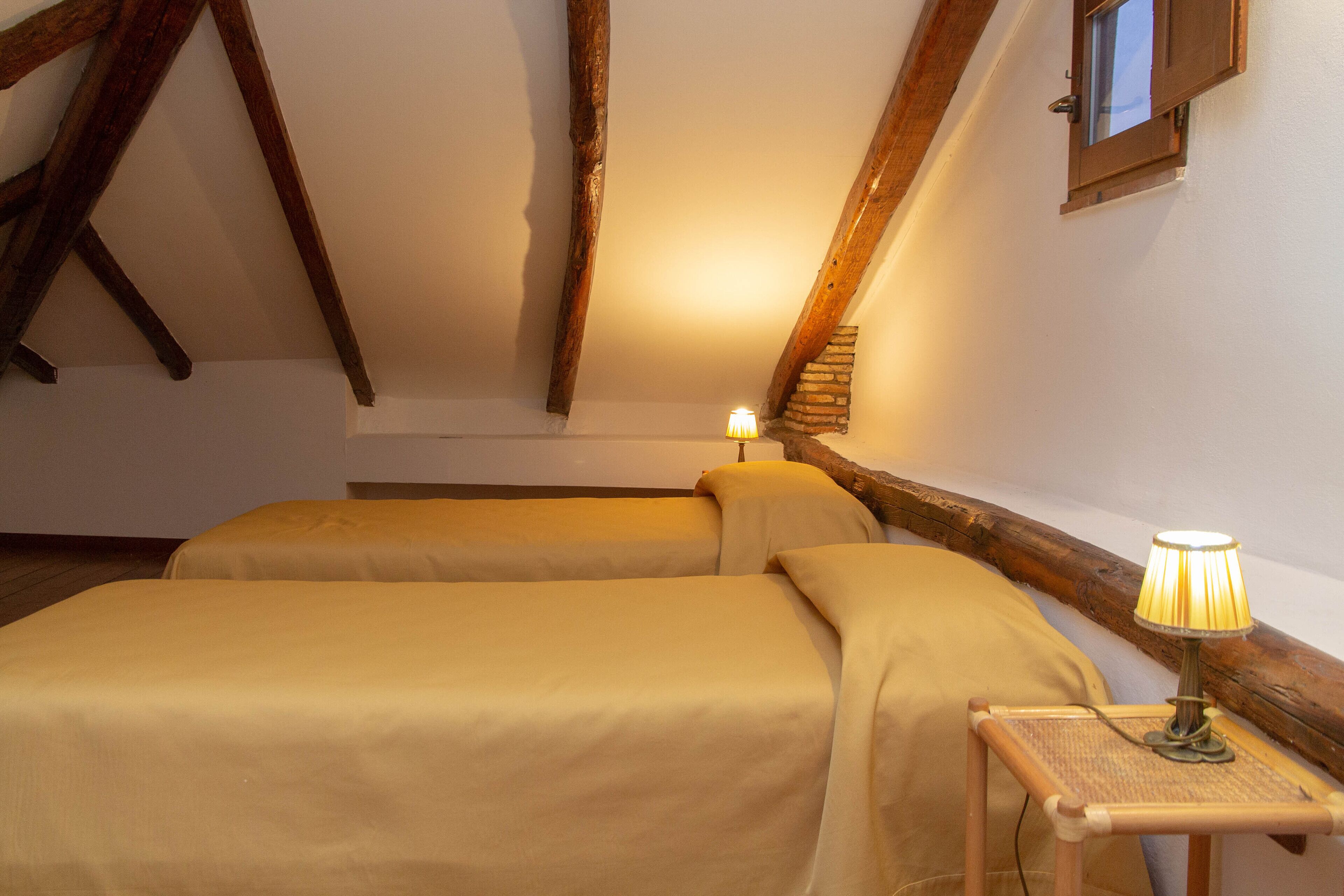 Une chambre avec deux lits, des poutres en bois, une petite fenêtre et une lampe de table.