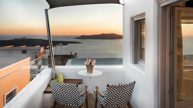 A balcony with a view of the sea at sunset, featuring two chairs with patterned cushions, a small table with a vase, and a glass railing. - Emfasis Villa (Santorini)