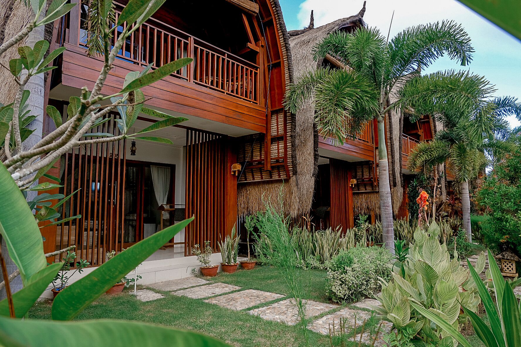 A wooden house with a balcony, surrounded by tropical plants and a stone pathway.