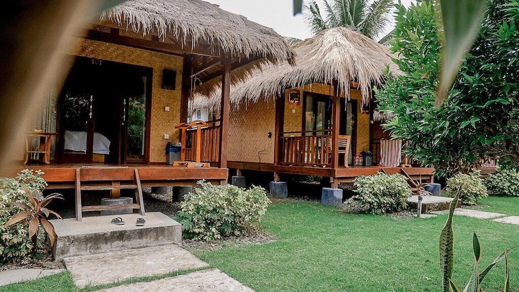 A bungalow-style building with thatched roofs, a wooden deck, and a well-maintained garden with a lawn and various plants.