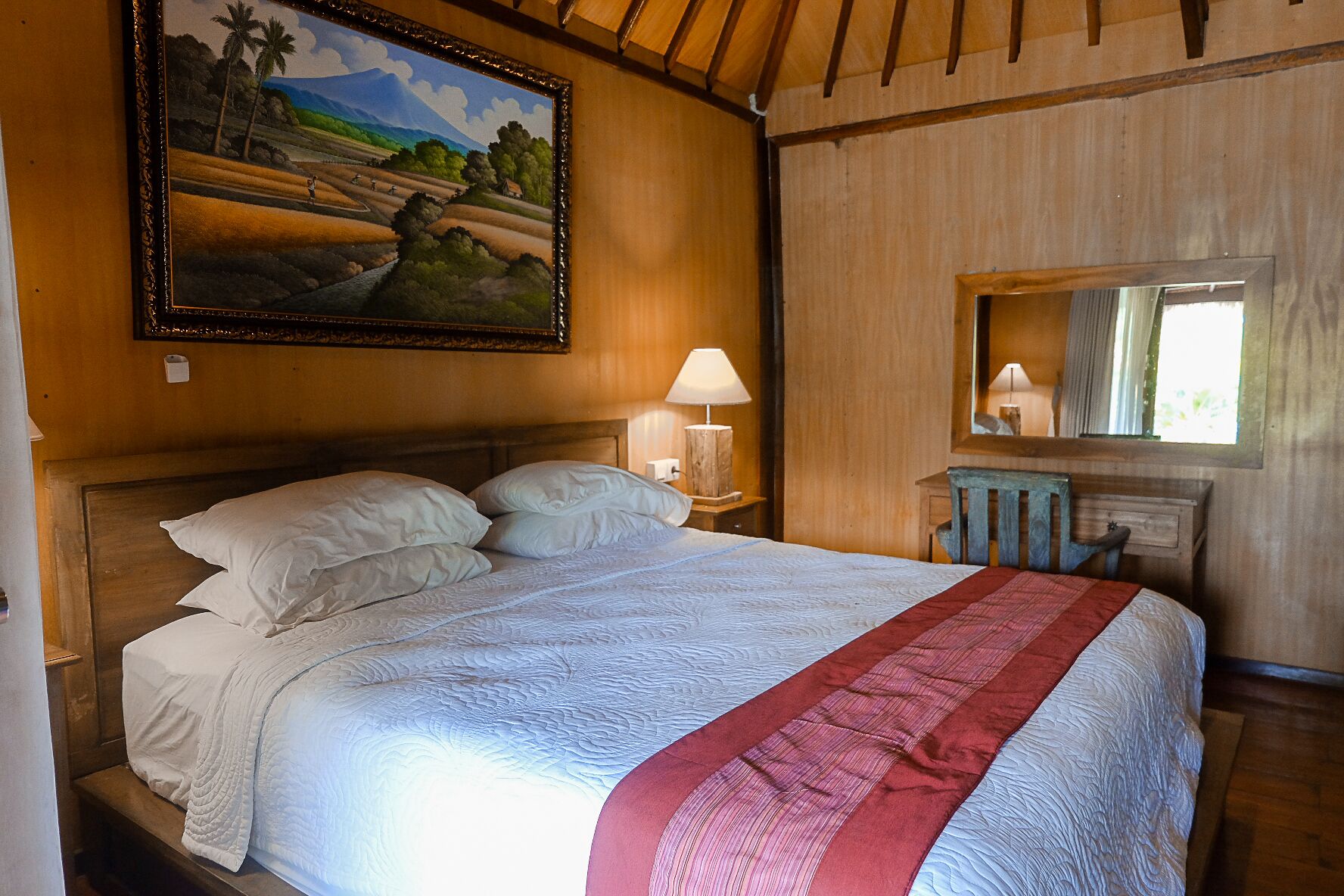 A bedroom with a large bed, a nightstand with a lamp, a wooden headboard, a painting on the wall, and a window with a view.