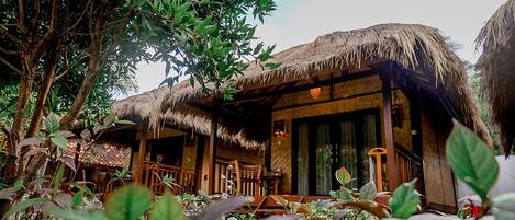 A rustic thatched-roof building with a wooden porch and large windows, surrounded by lush greenery.